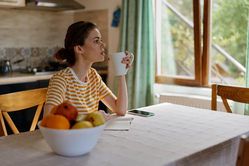A woman sits at a sunlit kitchen table, sipping from a white cup. A bowl of fruit sits nearby, striped tee, reflective mood. A quiet morning routine in a cozy home setting.