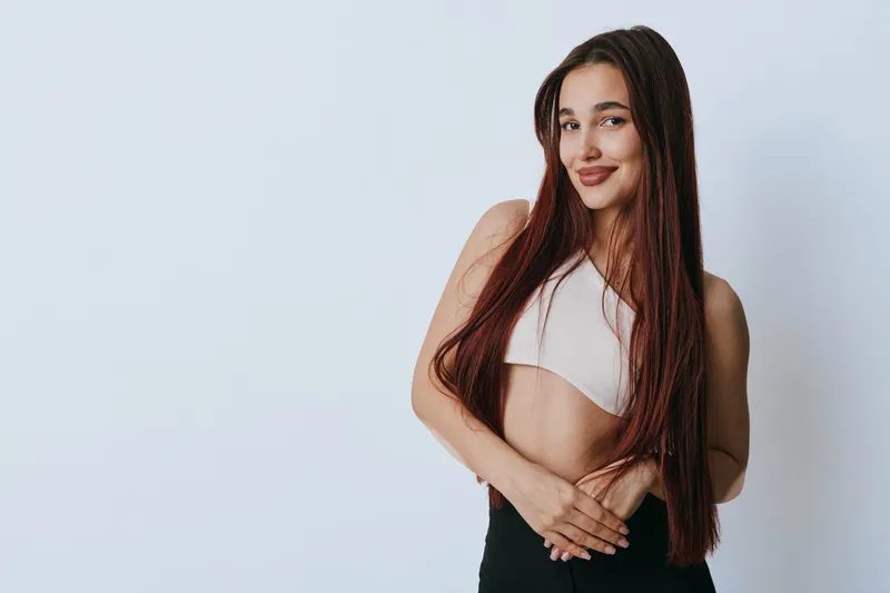 Young woman with long hair and braids posing in white crop top and black pants, smiling confidently against plain light background with hands crossed at waist.