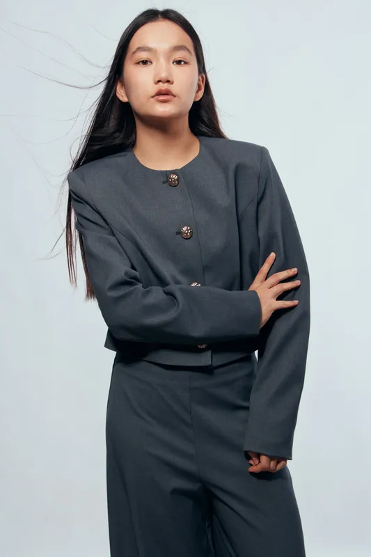 Young woman with long dark hair wearing stylish dark gray suit posing with folded arms on light background. Fashion, confidence, elegance, and modern business style in studio portrait.