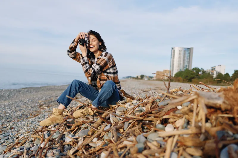 Beach scene with a smiling woman kneeling among pebbles posing with binoculars and camera, casual outdoor photography moment on the coast, breezy fashion and nature setting