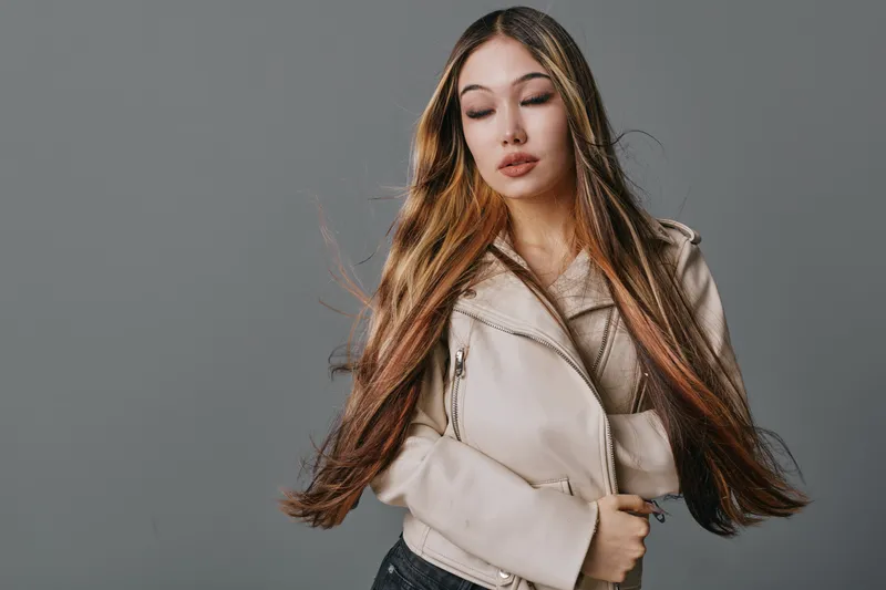 Young woman with long straight hair wearing beige leather jacket posing against gray background for fashion portrait and style concept with closed eyes expression.