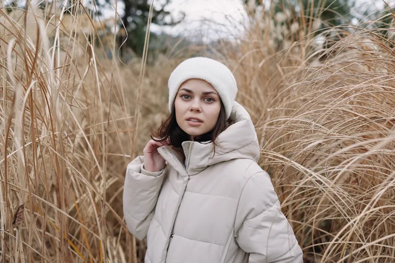 Winter scene with a woman in a light puffer coat and hat standing among tall grasses, conveying calm, candid fashion and outdoor serenity in soft natural light.