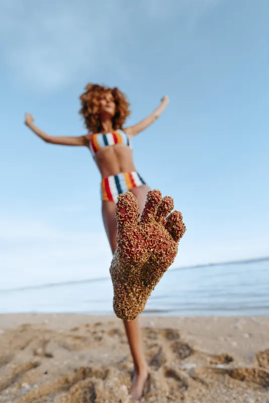 Joyful woman on beach in summer, jumping with arms up, striped swimsuit, heart shaped sand in foreground, warm sunlight, blue sky, calm sea waves, carefree mood, soft sea breeze today