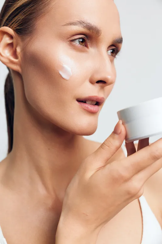 A closeup of a young woman applying cream on her cheek with a white jar in hand. The natural light highlights her healthy skin and focused expression in a minimal background.