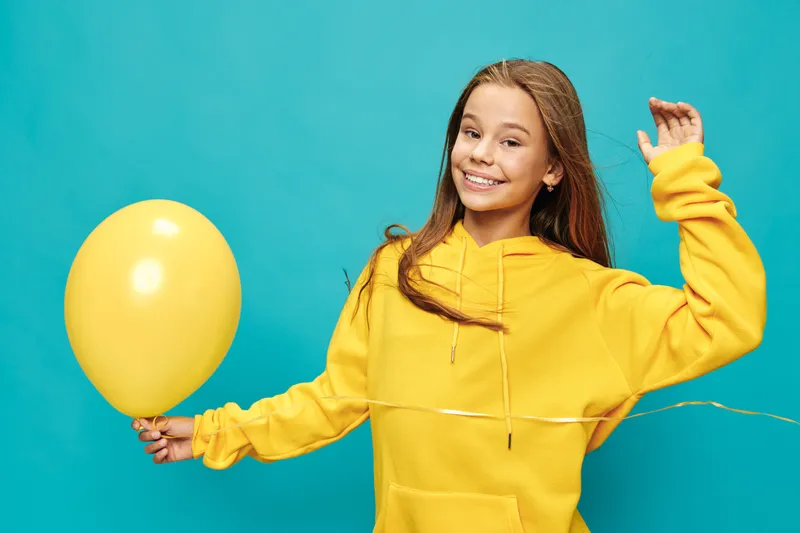 Girl smiling and holding yellow balloon on blue background wearing bright yellow hoodie for celebration and fun in studio. Happy child with long hair in casual clothing enjoying party moment.