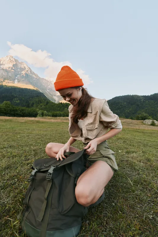 A woman outdoors in a bright field with an orange beanie bows over a backpack, preparing gear in a scenic mountain landscape for an adventurous lifestyle moment.