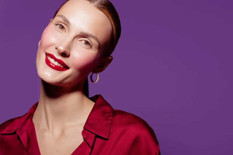 Smiling woman with red lipstick and hoop earrings poses against purple background. She wears a red satin blouse and has smooth skin with natural makeup. Concept of beauty and fashion.