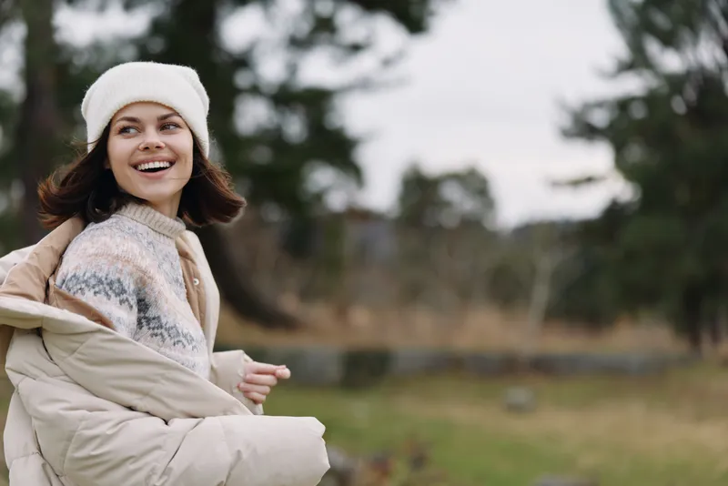 woman outdoor smiling portrait in nature, wearing a cozy sweater and beanie, sitting in a park during autumn, conveying relaxed mood, warmth and casual outdoor lifestyle in a serene setting