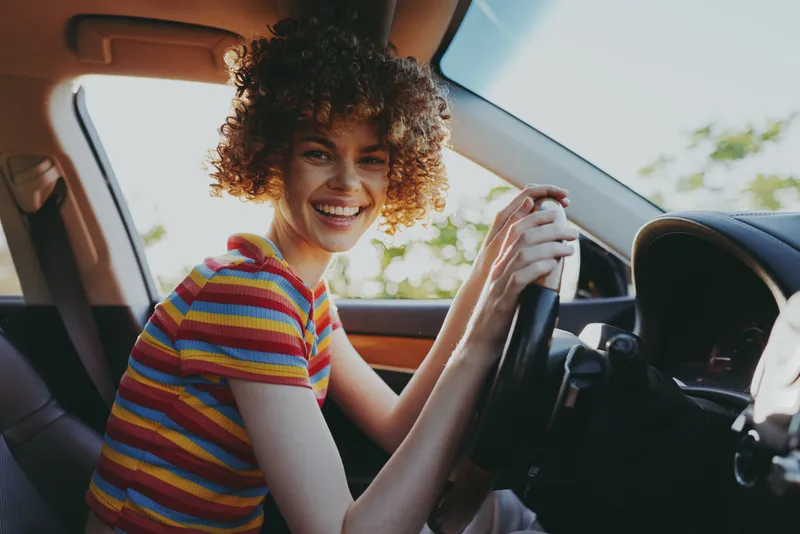 Candid woman inside a car, smiling at the wheel, daylight through the window, casual driving pose, retro color mood, warm interior tones, relaxed lifestyle moment