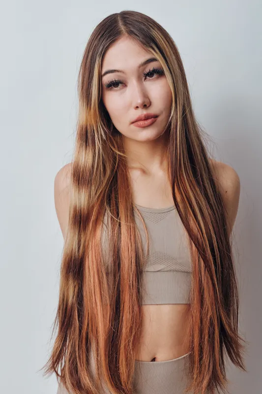 Young woman with long straight hair wearing beige sleeveless top against white background. Portrait of beauty model with natural makeup and confident expression standing indoors.