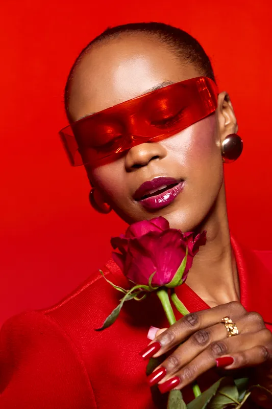 Close-up of an elegant dark skinned African woman wearing red sunglasses and blazer holding a red rose on a vibrant red background. Concept of beauty, fashion, and confidence.