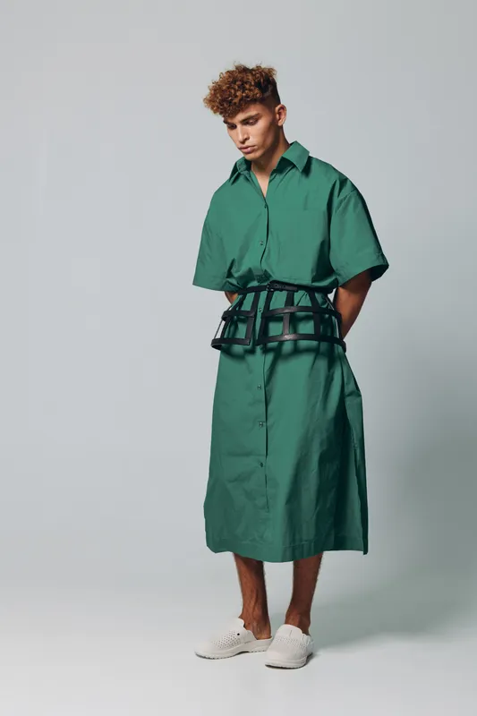 Young man with curly hair wearing green oversized shirt dress and white sneakers standing against plain background with hands behind back in casual fashion style.