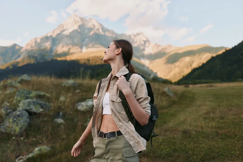 woman outdoors, hiking with backpack, mountain landscape, sunlit moment, relaxed posture, fresh air, natural colors, serene breeze, carefree lifestyle, scenic adventure