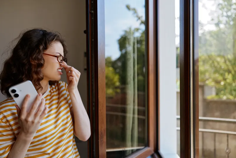 Casual woman with striped shirt stands by a sunny window checking her smartphone, relaxing at home. Natural light highlights a calm mood and everyday selfcare.