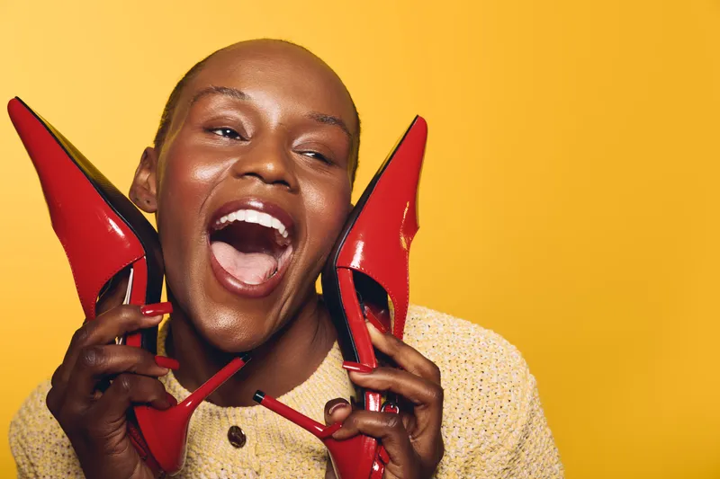 Happy African woman with dark skin laughing and holding red high heel shoes near her face on yellow background. Concept of fashion, joy, and beauty with vibrant colors and expression.