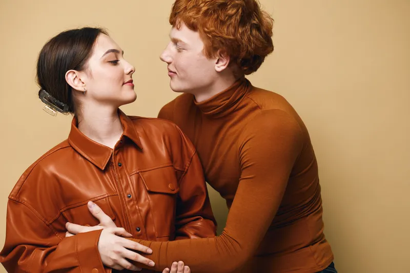 Couple, young man and woman embracing and looking at each other with affection and smile in studio with beige background and cozy autumn clothing.