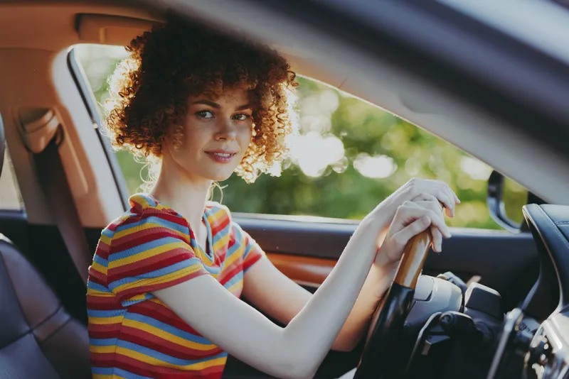 A woman driver sits behind the steering wheel in a sunny car, wearing a striped tee, smiling softly. A lifestyle portrait with retro film color and carefree road trip mood in bright light.