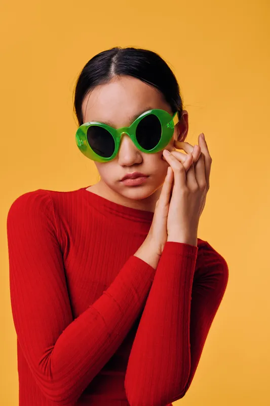 Woman wearing green round sunglasses and red long sleeve top posing with hands near face on orange background. Fashion portrait of young female model with minimal makeup and sleek hairstyle.