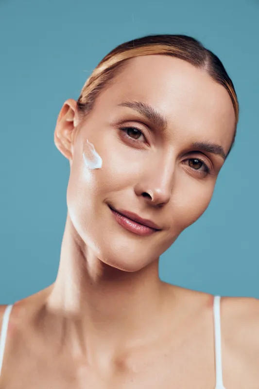 Young woman with clear skin applies moisturizing cream on face looks at camera with soft smile isolated on blue background natural beauty skincare concept.
