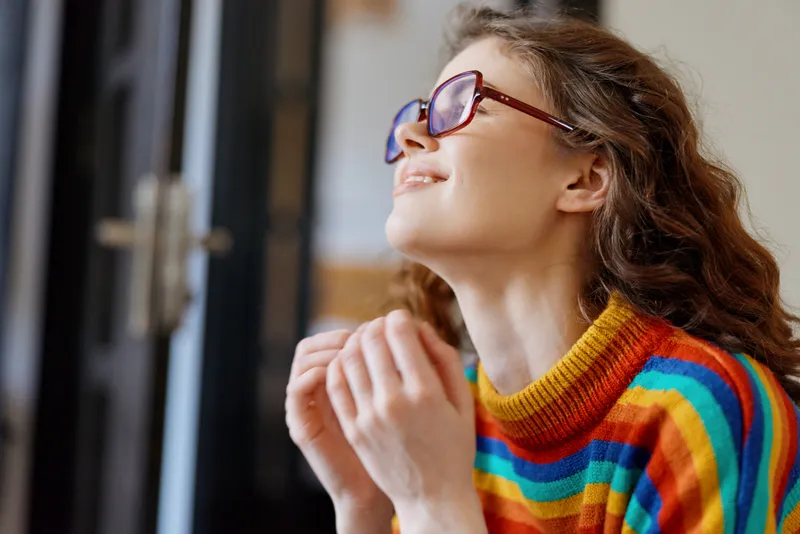 Woman in striped rainbow sweater wearing glasses smiles softly near a doorway, capturing a bright casual lifestyle moment in a cozy home setting, with a warm, relaxed mood.