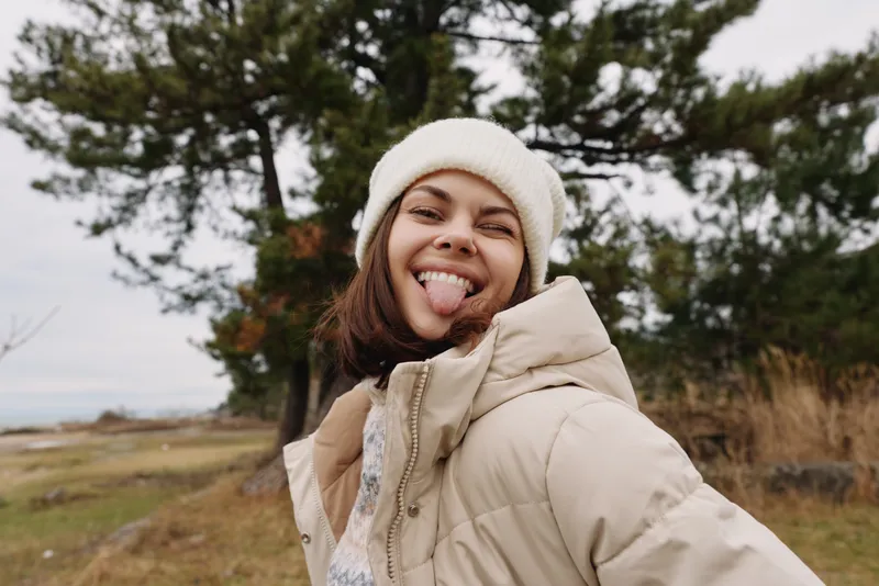 Playful woman outdoors in a warm beige jacket and white beanie, sticking out tongue with a joyful expression, set against a grassy field and pine trees under a cloudy sky.