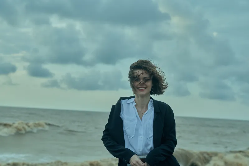 Woman in blazer and white shirt stands on a windy beach by the sea, smiling under a cloudy sky, showcasing professional style and outdoor mood with coastal atmosphere for lifestyle and work content