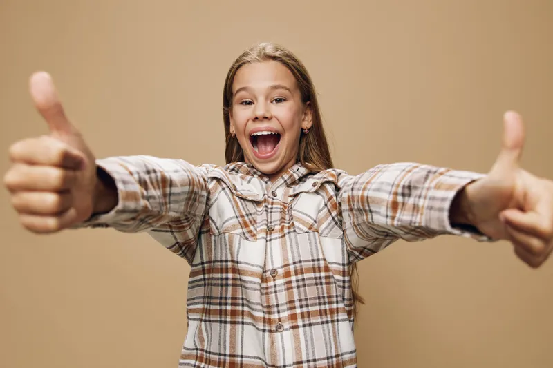 Girl teenager in plaid shirt smiling and showing thumbs up with both hands, emotion of happiness and approval, isolated on beige background for positive expression.