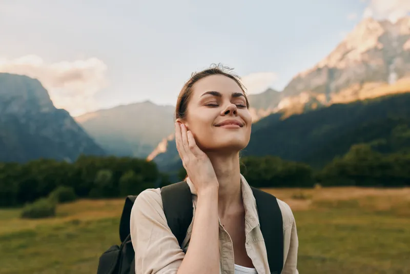 Young woman with closed eyes relishes a sunlit mountain breeze during a peaceful outdoor hike, backpack resting securely as distant peaks glow softly and clear blue sky frames her calm smile.