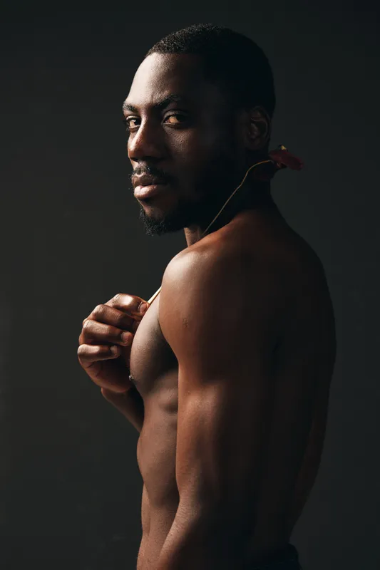 African man with dark skin and muscular body posing shirtless with flower on shoulder in creative conceptual visual style against dark background. Artistic portrait with moody lighting.