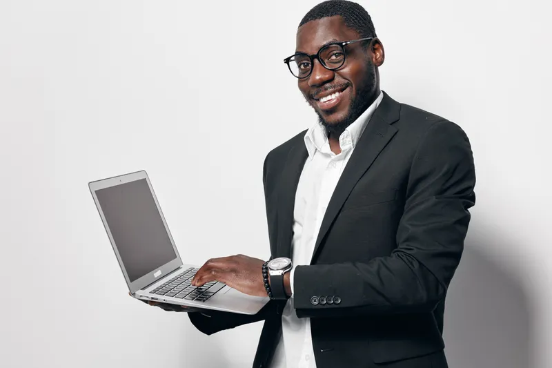 African American man with dark skin wearing glasses and black suit holding laptop smiling at camera on white background. Business professional, technology and communication concept.