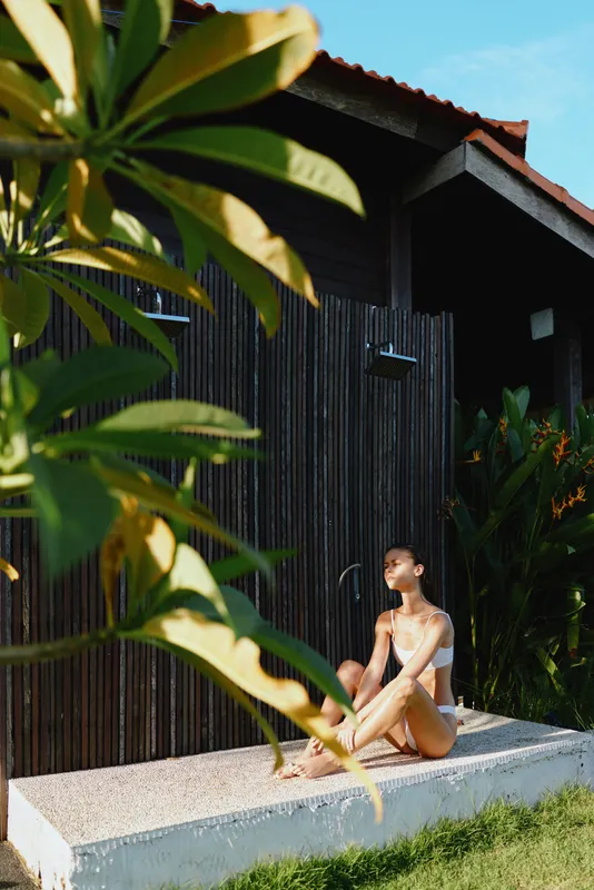 Young woman in white swimsuit sitting on concrete ledge near tropical plants and dark wooden house. Bright sunlight highlights summer lifestyle, relaxation, and outdoor leisure mood.