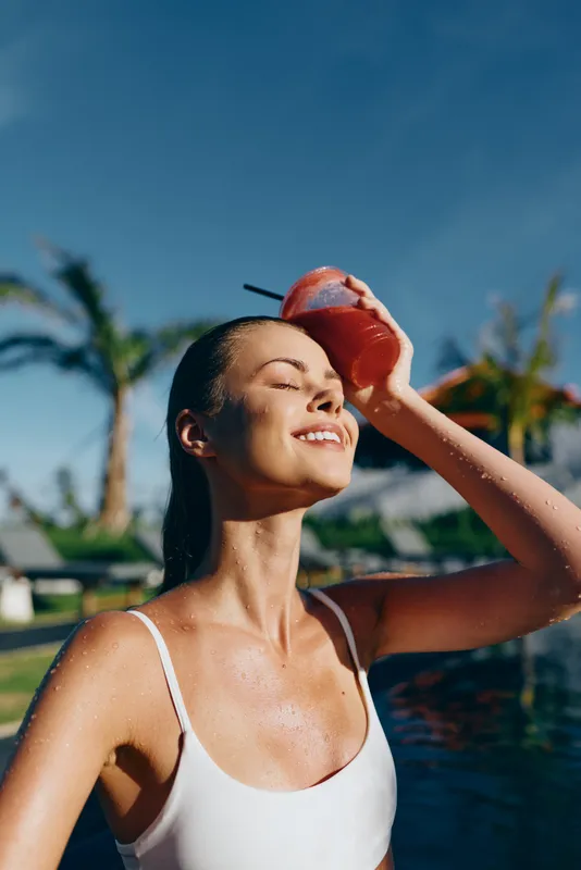 A smiling woman in a white swimsuit enjoys sunshine beside a pool, tilting her head as she drinks from a coconut. A bright, carefree moment capturing relaxed summer lifestyle.