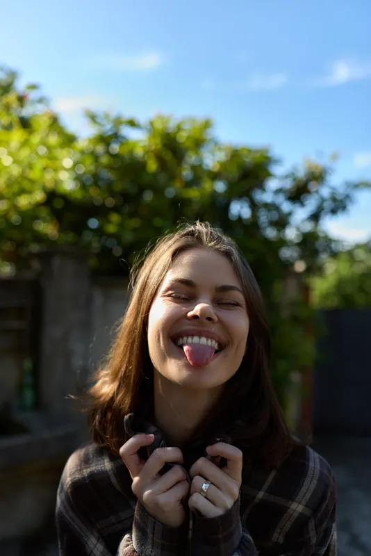 Cheerful woman outdoors, smiling with playful energy, sunlight warming the sky, candid portrait capturing a carefree lifestyle in a garden setting