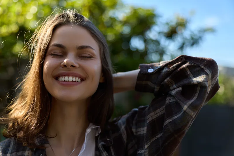 A cheerful woman outdoors basks in sunlight, wearing a plaid shirt and white top, eyes closed, smiling softly. A lifestyle portrait capturing relaxed confidence and carefree moment in a bright garden