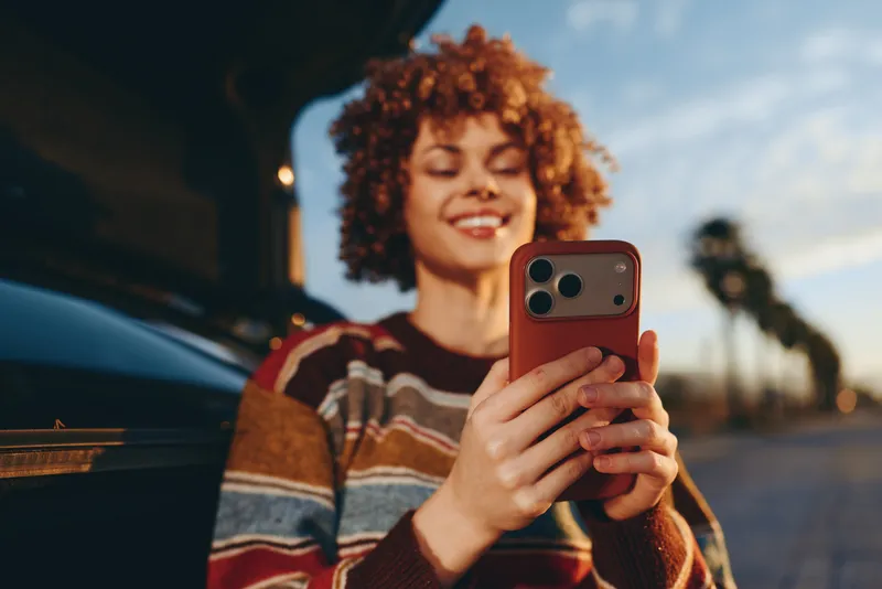 Woman with curly hair holds a smartphone and takes a selfie beside a car on the street, lifestyle casual outdoor moment, warm evening light, relaxed smile, friendly gaze