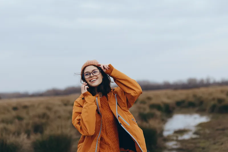 Woman lifestyle outdoors countryside portrait in autumn, smiling young woman in brown coat and hat, adjusting headwear while holding scarf, grassy field and cloudy sky, soft light