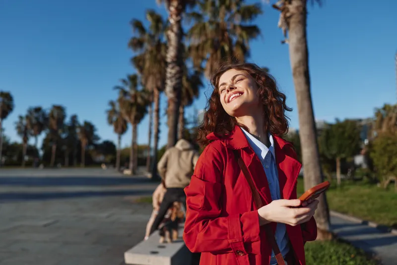 Smiling woman with curly hair wearing a red coat outdoors in a sunny park with palm trees. Fashionable, upbeat lifestyle moment captured in warm natural light.