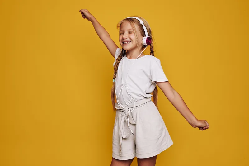 Happy girl with headphones dances with closed eyes in casual clothes on yellow background studio.