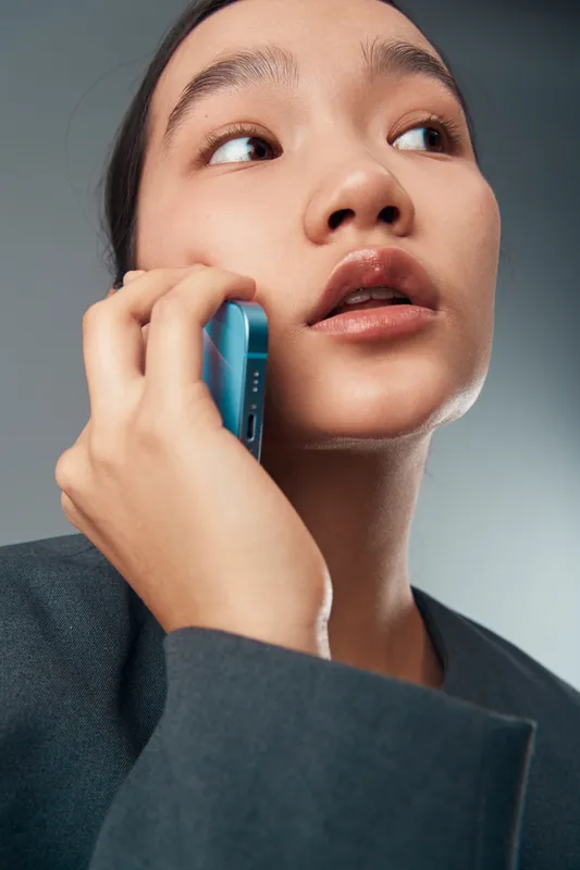 Young woman holding smartphone close to ear making call or talking with focused expression wearing dark jacket against neutral background in closeup portrait style.