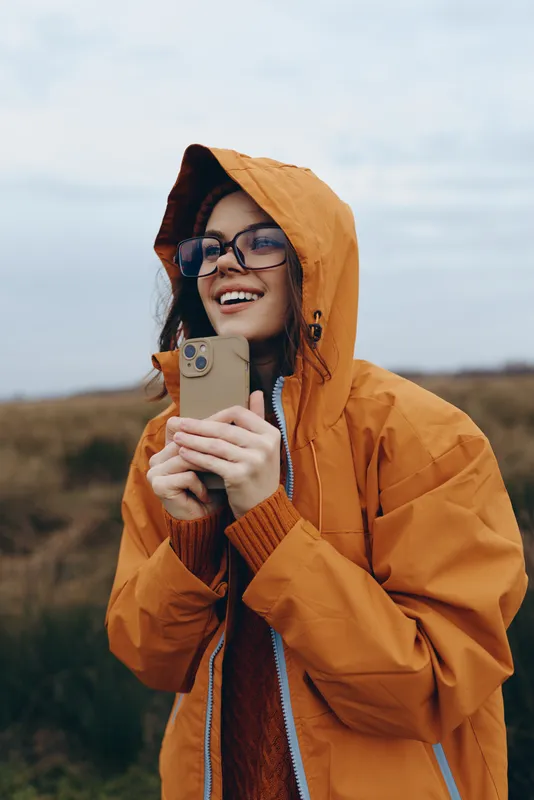 Woman lifestyle outdoors, hooded jacket, smiling, holding a smartphone, cheerful portrait in countryside field, cloudy sky background, natural light, casual travel concept, relaxed communication