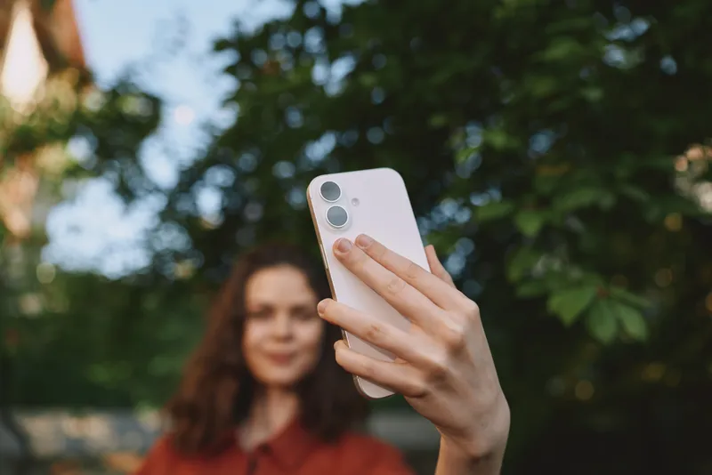 A smiling woman takes a selfie outdoors with a smartphone, casual fashion, natural light, trees in the background, capturing a bright everyday lifestyle moment