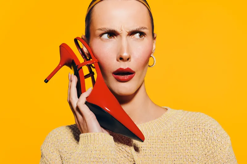 Confused woman in beige sweater holds red high heel shoe near ear on yellow background. Concept for fashion, style, and unusual communication or creativity.