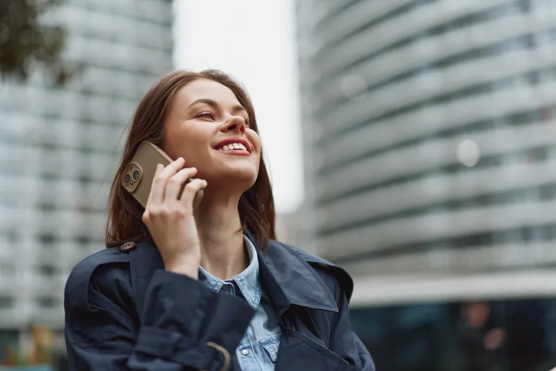 Young woman in a navy coat chats on a smartphone in an urban plaza, smiling as tall buildings rise behind, capturing a candid city moment and lively energy.