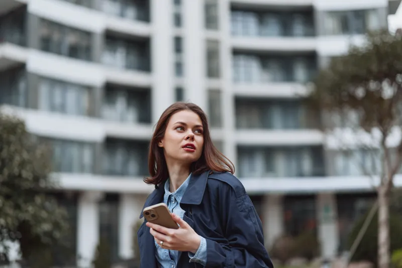 A young woman in a denim jacket stands outdoors near a modern apartment building, holding a smartphone and looking up with curiosity and thoughtful interest.