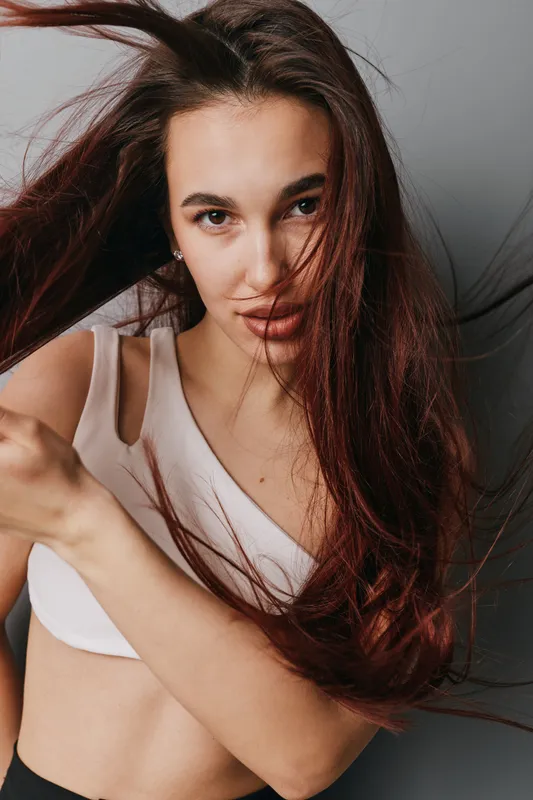 Young woman with long brown hair wearing white top posing indoors with hair flowing, looking at camera with confident expression and natural makeup on gray background.