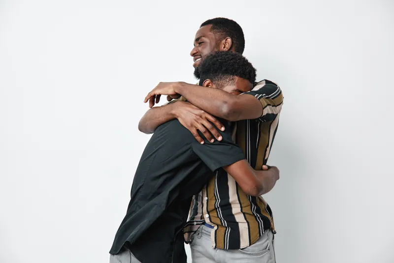 Two African American men with dark skin hugging each other in casual clothes for friendship, support, and connection on white background. Emotional embrace and male bonding.