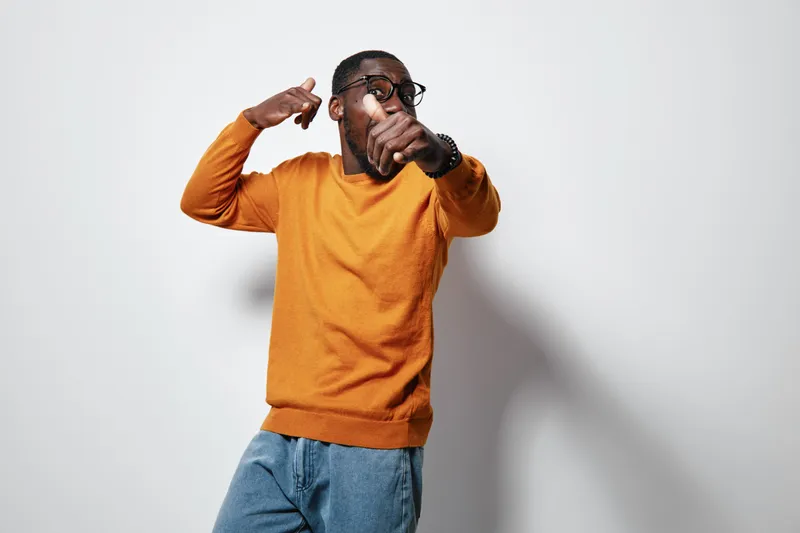 African American man with dark skin wearing orange sweater and glasses posing with confident gesture on white background. Casual style and modern look.