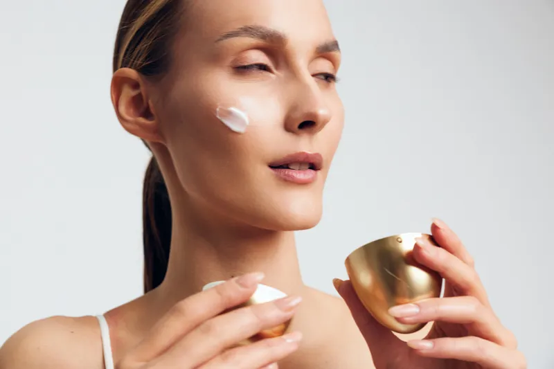 Young woman applying white cream to her cheek in a clean studio, showcasing skincare routine and self care with soft, warm lighting and a calm mood.