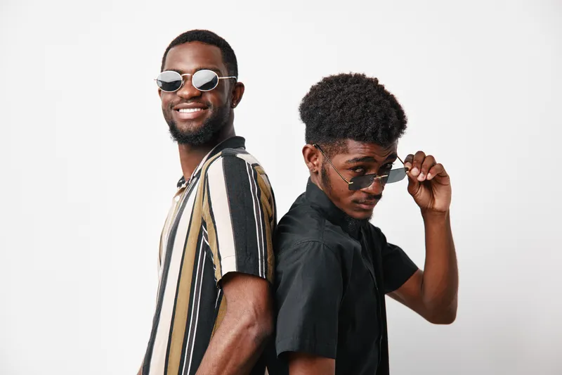 Two young black men with dark skin posing back to back wearing sunglasses and casual shirts, smiling and confident in studio with white background for lifestyle and friendship.
