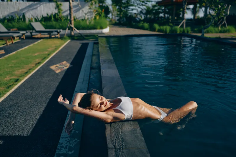 A young woman in white bikini relaxes on the edge of an outdoor pool with her arms stretched back. The sunny day highlights her lifestyle and leisure time by the water.
